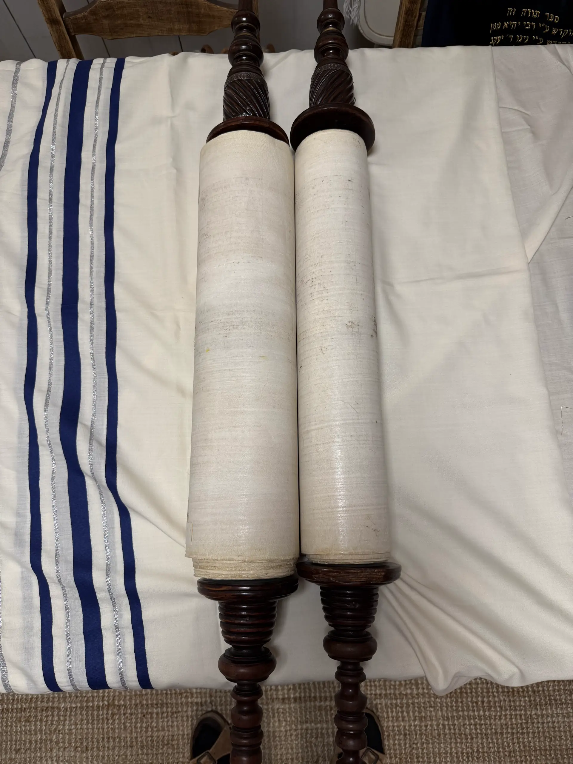 A Sefer Torah rolled on its atzei chayim, resting on a tallit. The embroidered mantle carries a dedication.